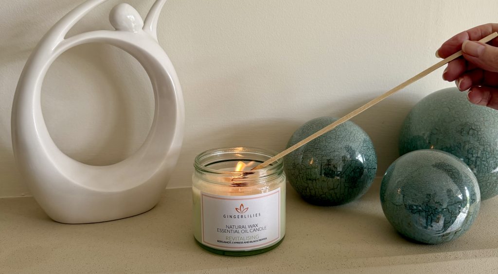 A hand using a long wooden match to light a Gingerlilies 'Revitalising' natural wax essential oil candle on a shelf, flanked by a white abstract sculpture and green crackle-glazed decorative spheres.
