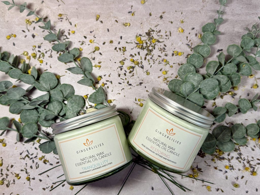 An overhead flat lay showing two Gingerlilies natural wax essential oil candles—'Tranquillity' and 'Restoring'—nestled among green eucalyptus branches, scattered dried chamomile, and lavender blossoms on a textured stone surface.
