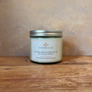 Gingerlilies 'Restoring' Essential Oil Candle with a silver lid, featuring a white label with rose geranium, vetiver, and patchouli scents, displayed on rustic wood flooring.
