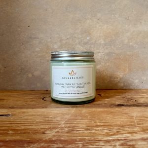 Gingerlilies 'Restoring' Essential Oil Candle with a silver lid, featuring a white label with rose geranium, vetiver, and patchouli scents, displayed on rustic wood flooring.