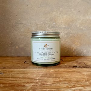 Gingerlilies 'Revitalising' Essential Oil Candle with a silver screw-top lid, featuring a white label with bergamot, cypress, and black pepper ingredients, displayed on rustic wood flooring.